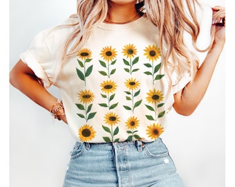 May include: A white t-shirt with a pattern of yellow sunflowers on green stems. The shirt is being worn by a person with long blonde hair.