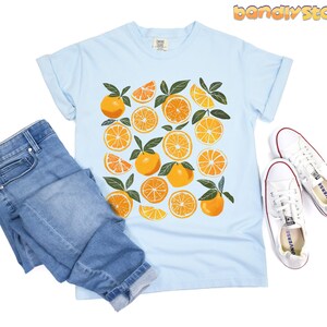May include: A light blue t-shirt with a graphic print of orange slices and leaves. The shirt is paired with blue jeans and white sneakers.