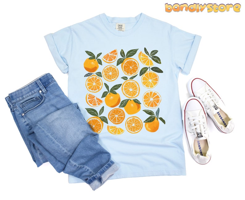 May include: A light blue t-shirt with a graphic print of orange slices and leaves. The shirt is paired with blue jeans and white sneakers.