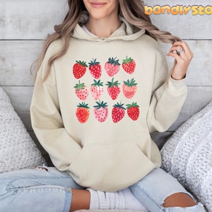 Pode incluir: Um moletom de cor creme com um gr&aacute;fico de morangos vermelhos e rosados. Os morangos est&atilde;o dispostos em uma grade.