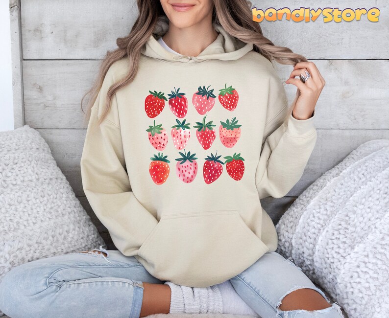 Pode incluir: Um moletom de cor creme com um gr&aacute;fico de morangos vermelhos e rosados. Os morangos est&atilde;o dispostos em uma grade.