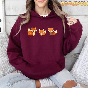 May include: A maroon hoodie with a graphic of three foxes. The foxes are orange with white accents. The hoodie has a drawstring hood and a kangaroo pocket.