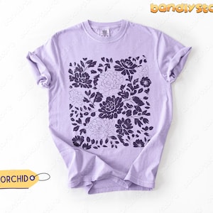 Op de afbeelding: Een lichtpaars t-shirt met een donkerpaars bloemenpatroon. Het ontwerp toont verschillende bloemen en bladeren gerangschikt in een vierkant patroon. Het woord "ORCHID" staat op een geel label dat aan het shirt is bevestigd.