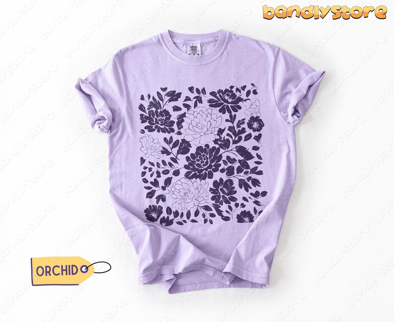 Op de afbeelding: Een lichtpaars t-shirt met een donkerpaars bloemenpatroon. Het ontwerp toont verschillende bloemen en bladeren gerangschikt in een vierkant patroon. Het woord "ORCHID" staat op een geel label dat aan het shirt is bevestigd.