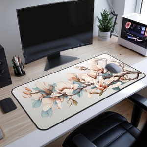 May include: A rectangular mouse mat with a floral design. It features magnolia blossoms in pink, peach, and cream, set against a light grey background. The blossoms are arranged along a brown branch with light blue leaves. A grey mouse rests on the mat.