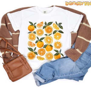 May include: White t-shirt with a graphic print of orange slices. The shirt is paired with brown and white striped sweater and blue jeans.