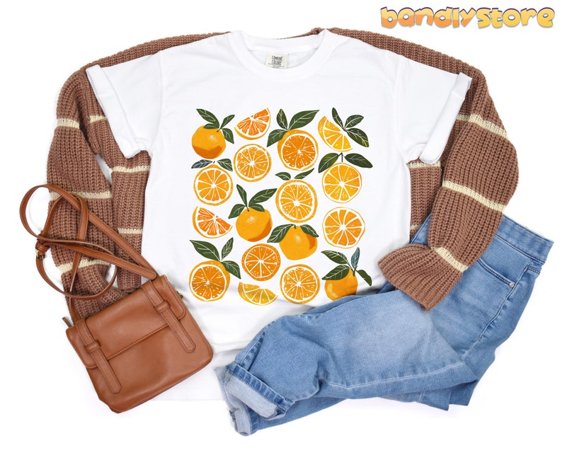 May include: White t-shirt with a graphic print of orange slices. The shirt is paired with brown and white striped sweater and blue jeans.