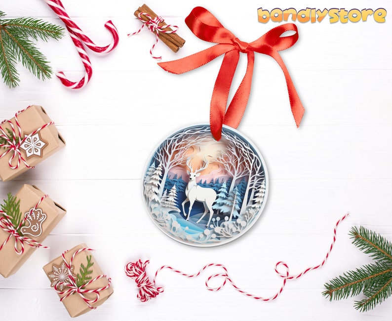 May include: A white papercraft deer ornament with a blue and white winter forest scene. The ornament is hanging from a red ribbon and is surrounded by Christmas decorations.