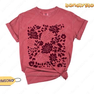 Op de afbeelding: Een stoffig roze t-shirt met een donker bordeauxrood bloemmotief. Het bloemmotief is een vierkante opstelling van verschillende bloemen en bladeren. Het shirt heeft opgerolde mouwen en een knoop aan de onderkant. Het label zegt "CRIMSONG".
