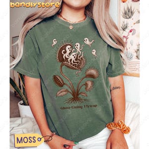 May include: Moss green t-shirt featuring a detailed illustration of a Venus flytrap with ghosts and the text "Ghost-Eating Flytrap." The design is in shades of brown and white. The shirt has short sleeves and a crew neck.