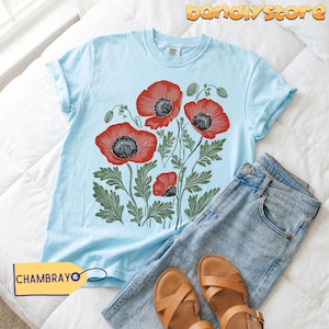 May include: A light blue t-shirt with a red poppy flower design and green leaves. The shirt is styled with blue jeans and brown sandals. The tag reads CHAMBRAYO.