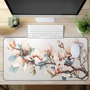 May include: A rectangular mouse mat with a floral design. Large, light pink magnolia blossoms and blue-green leaves are on a light grey background. The blossoms are on a brown branch. A white mouse and keyboard are on the mat.