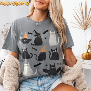 May include: A gray t-shirt with a black and white print of cats wearing witch hats and holding brooms. The cats are surrounded by bats and stars. The text "bandystore" is printed in the top left corner of the shirt.