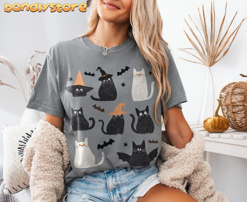 May include: A gray t-shirt with a black and white print of cats wearing witch hats and holding brooms. The cats are surrounded by bats and stars. The text "bandystore" is printed in the top left corner of the shirt.