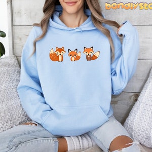 May include: A light blue hoodie with a graphic of three cartoon foxes. The foxes are orange and white with black eyes and noses. The hoodie has a drawstring hood and a kangaroo pocket.