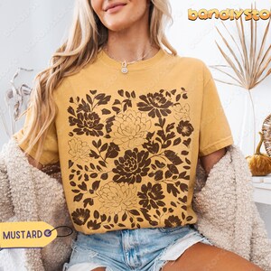 Op de afbeelding: Mosterdkleurig T-shirt met een bruin bloemenpatroon. Het T-shirt heeft een ronde hals en korte mouwen. De tekst "MUSTARD" staat op een geel label. Het model draagt een blauwe spijkerbroekshort en een cr&egrave;me vest.