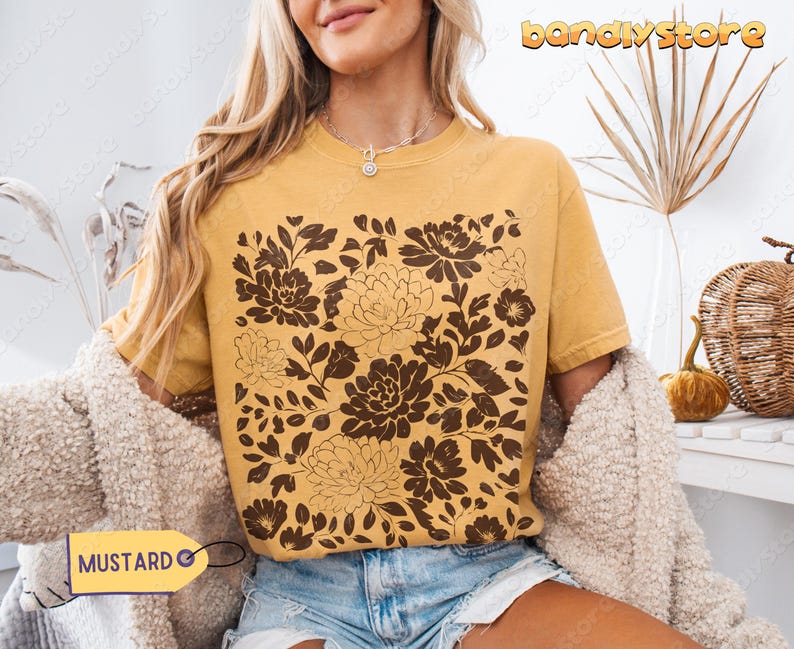 Op de afbeelding: Mosterdkleurig T-shirt met een bruin bloemenpatroon. Het T-shirt heeft een ronde hals en korte mouwen. De tekst "MUSTARD" staat op een geel label. Het model draagt een blauwe spijkerbroekshort en een cr&egrave;me vest.