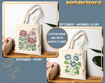 May include: Two white canvas tote bags with custom names and birth month flowers. The bag on the left has a pink and green floral design and the text "Alice" and "NOVEMBER - PEONY". The bag on the right has a blue and green floral design and the text "Phoebe" and "SEPTEMBER - MORNING GLORY".