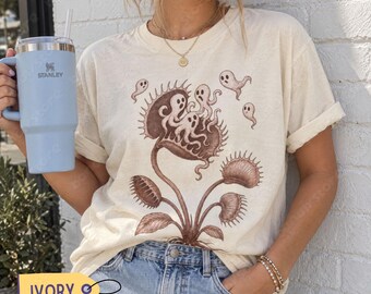 May include: An ivory t-shirt featuring a brown graphic print of a Venus flytrap plant with multiple small ghosts emerging from its leaves. The shirt is paired with blue denim jeans and a light blue Stanley tumbler with a straw.