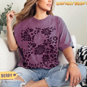 Woman in a muted purple (berry) tee featuring deep plum linocut flowers. The shirt has a cozy, relaxed cottagecore feel.