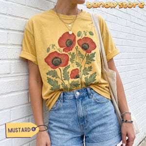 May include: Mustard yellow t-shirt with a red poppy floral print and green leaves. The shirt is worn with denim shorts. The word "MUSTARD" is on a yellow tag. The sleeves are rolled up.