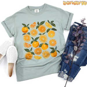 May include: A light blue t-shirt with a graphic print of orange slices on a white background. The shirt has a crew neck and short sleeves.