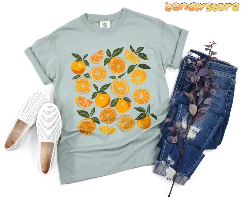 May include: A light blue t-shirt with a graphic print of orange slices on a white background. The shirt has a crew neck and short sleeves.