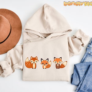 May include: A beige hoodie with a graphic of three orange foxes. The foxes are facing forward with their tails curled behind them. The hoodie is laid out on a white surface with a brown hat and blue jeans.