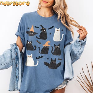 May include: A blue t-shirt with a graphic print of black and white cats wearing witch hats. The cats are surrounded by bats and stars.