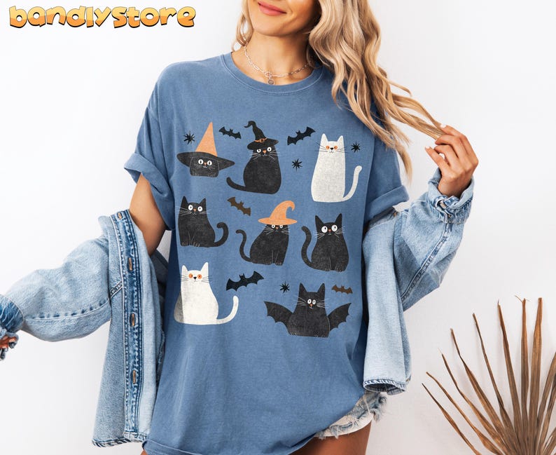 May include: A blue t-shirt with a graphic print of black and white cats wearing witch hats. The cats are surrounded by bats and stars.