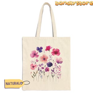 May include: A natural canvas tote bag with a design of watercolor pink, purple, and white flowers. The bag has a natural fabric handle.