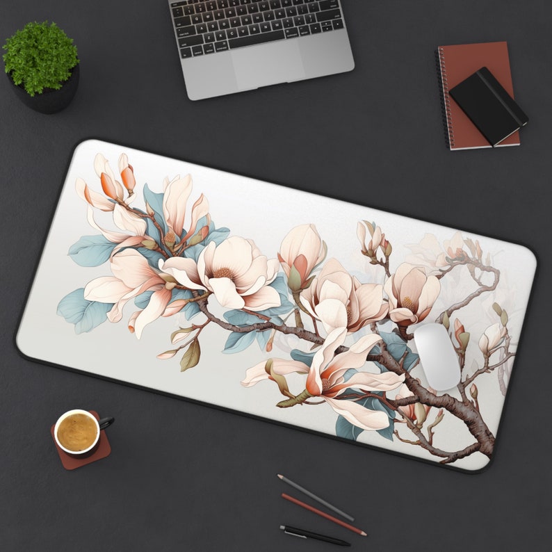 May include: A rectangular mouse mat with a floral design. It features large, light pink magnolia blossoms and blue-green leaves on a white background. The blossoms are set against a light grey backdrop, with a black border.