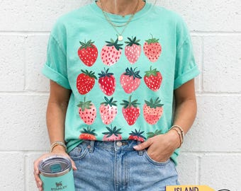 May include: A light green short-sleeve t-shirt featuring a repeating pattern of red strawberries. The shirt is paired with blue denim jeans and a light green insulated tumbler. The text bandlystore is visible in the top left corner of the image.