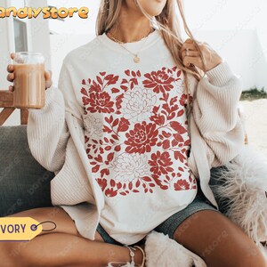 Woman wearing an ivory Comfort Colors tee with reddish-brown linocut-style flowers and leaves. The design has a handmade vintage stamp effect.