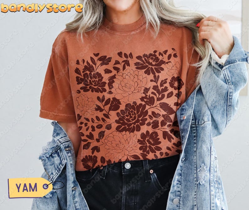 Woman in a rich yam orange tee showcasing a rustic dark red linocut floral print, styled with a denim jacket and black jeans.
