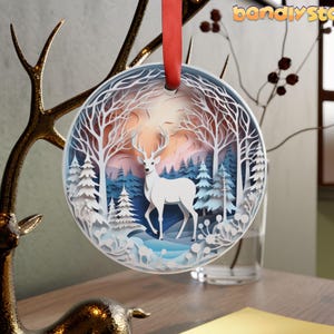 May include: White papercraft ornament featuring a deer standing in a snowy forest scene. The ornament is round and has a blue and white color scheme.