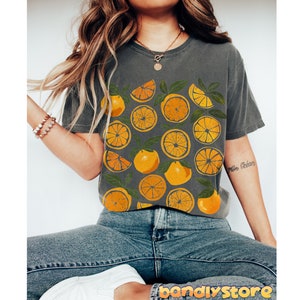 May include: A gray t-shirt with a graphic print of orange slices. The shirt is worn by a person with long brown hair and is paired with blue jeans.
