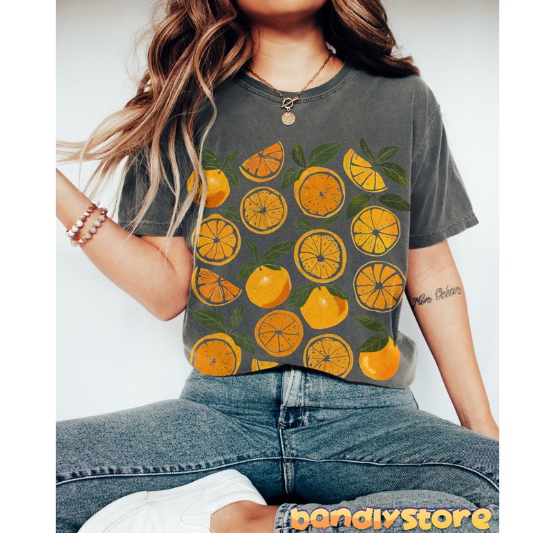 May include: A gray t-shirt with a graphic print of orange slices. The shirt is worn by a person with long brown hair and is paired with blue jeans.