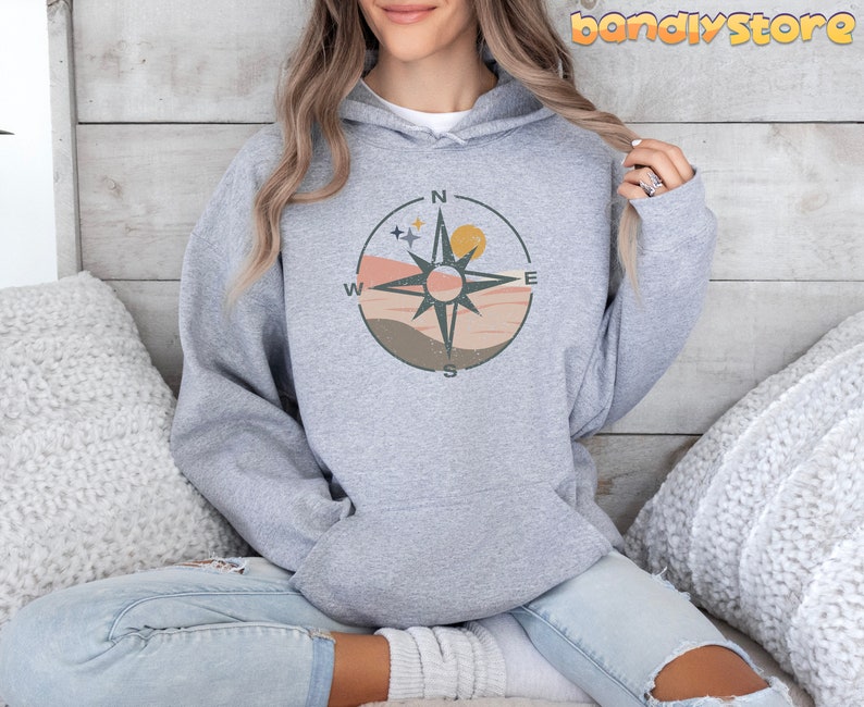May include: A gray hoodie with a compass design featuring a sunset, stars, and the letters N, E, S, and W. The compass is surrounded by a brown, pink, and orange background.
