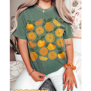 May include: A green t-shirt with a graphic print of yellow oranges and leaves. The shirt has a crew neck and short sleeves.