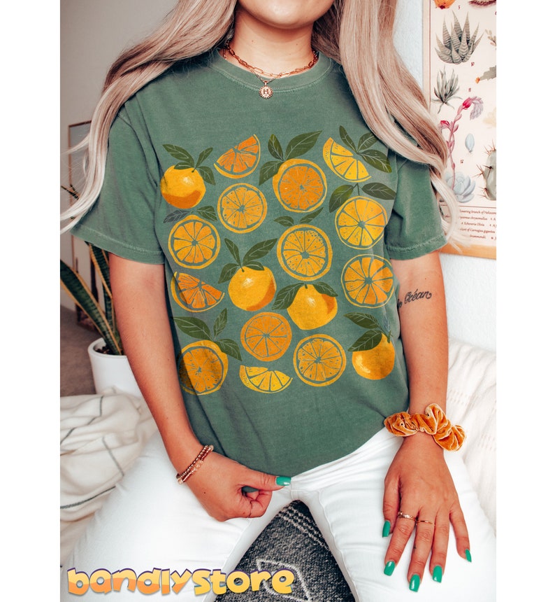 May include: A green t-shirt with a graphic print of yellow oranges and leaves. The shirt has a crew neck and short sleeves.