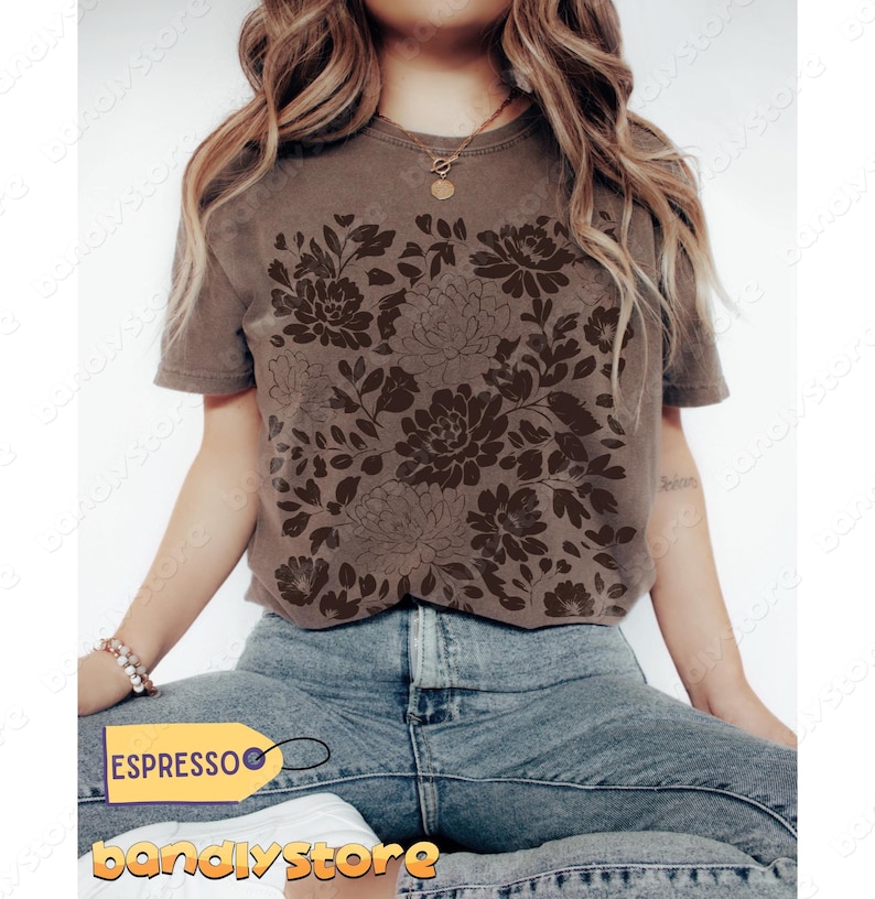 Woman wearing an espresso-colored Comfort Colors t-shirt featuring a large monochromatic linocut-style floral print across the front. The design showcases layered peonies and leafy foliage in a slightly darker tone for a subtle, textured look. She’s seated cross-legged in high-waisted light-wash jeans, accessorized with a gold pendant necklace and bracelet, against a minimal white background with soft natural lighting.