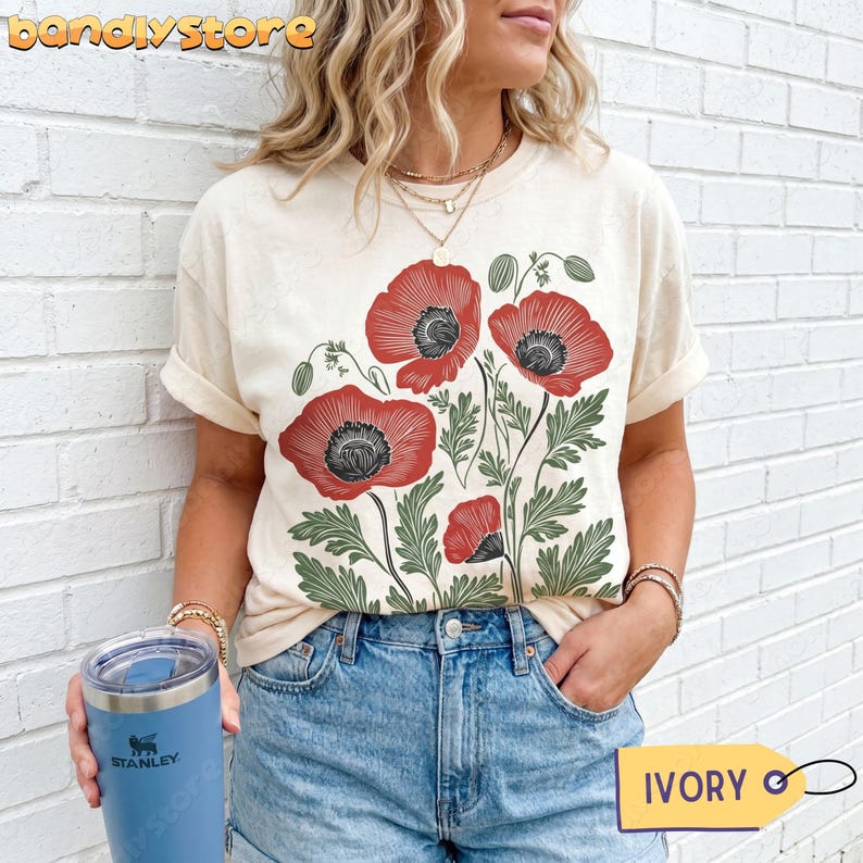 May include: Cream-coloured t-shirt with a red poppy floral design. The graphic features detailed red poppies with black centres and green leaves. The shirt has rolled-up sleeves and is paired with denim shorts. The word "IVORY" is on a tag.