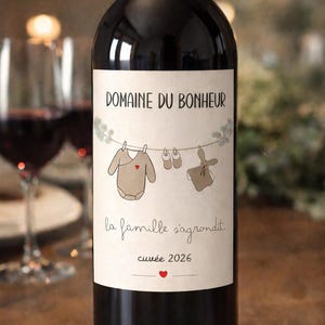 Peut inclure: Bouteille de vin rouge avec une étiquette indiquant "Domaine du Bonheur". L'étiquette présente une corde à linge avec des vêtements de bébé, des chaussures et un jouet. Le texte "la famille s'agrandit" et "cuvée 2026" sont également présents.