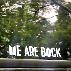 Puede incluir: Calcomanía de vinilo blanco con el texto "WE ARE BACK" en una fuente en negrita y sin serifa. La calcomanía está adherida a una superficie oscura, con un fondo borroso de follaje verde.