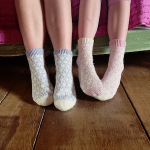May include: Two pairs of socks with a white and light purple geometric pattern. The socks are worn on the feet of two people sitting on a bed.