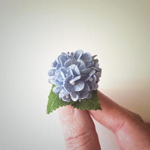 Op de afbeelding: Een gehäkelde blauwe hortensia bloem met een groen stoffen blad.