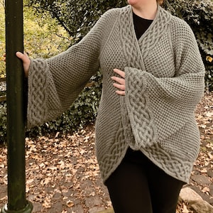 Op de afbeelding: Een grijze gebreide cardigan met een kabelbreipatroon en lange mouwen. De cardigan heeft een shawlkraag en een wikkelgesp.