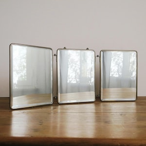 Op de afbeelding: Drie rechthoekige spiegels met een metalen frame staan op een houten oppervlak. De spiegels zijn scharnierend en verstelbaar. Het glas reflecteert de omgeving, wat een vintage esthetiek creëert.