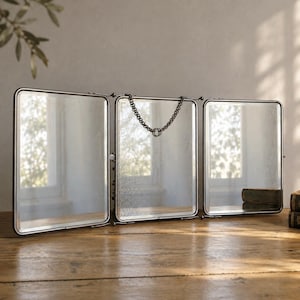 Può includere: Uno specchio pieghevole a tre pannelli con una cornice in metallo scuro. Il pannello centrale presenta una catena metallica decorativa appesa nella parte superiore. Le superfici in vetro hanno una finitura riflettente leggermente invecchiata e le cerniere consentono un posizionamento regolabile.